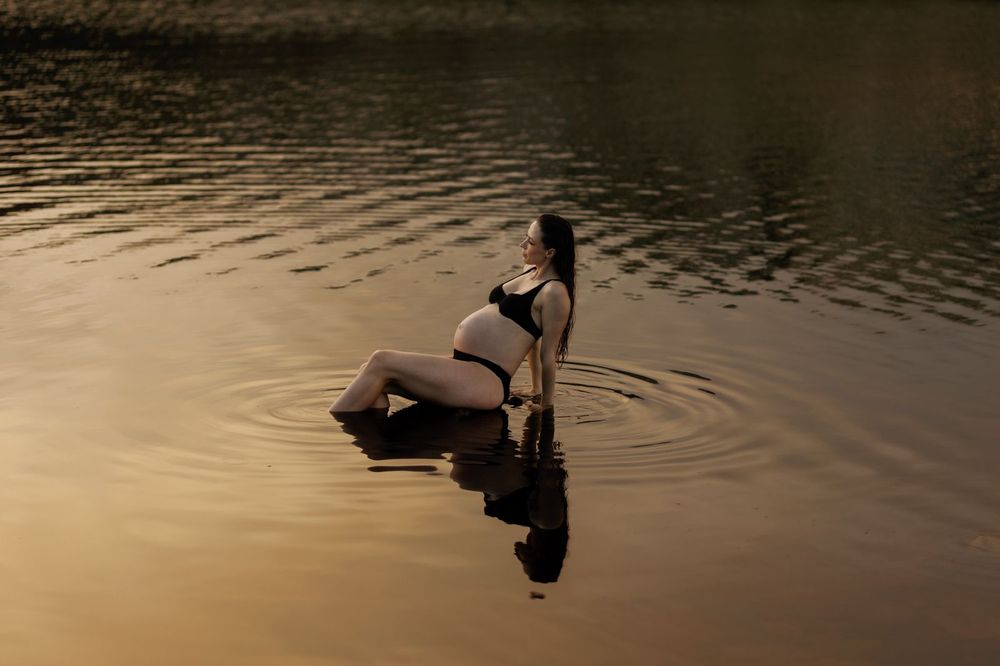 Pregnant woman in black bikini sitting peacefully in calm lake water at golden sunset, creating gentle ripples.