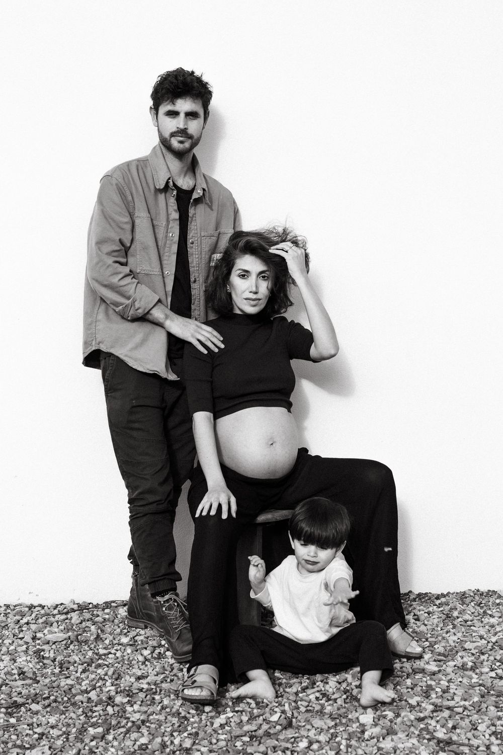 Black and white family portrait of pregnant woman, man, and toddler posing against white wall.