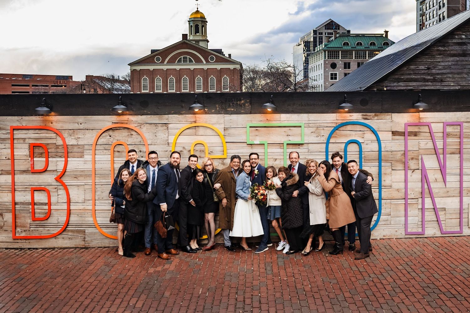 Bride and groom surrounded by their full wedding party and family in front of a colorful Boston sign in City Hall Plaza.