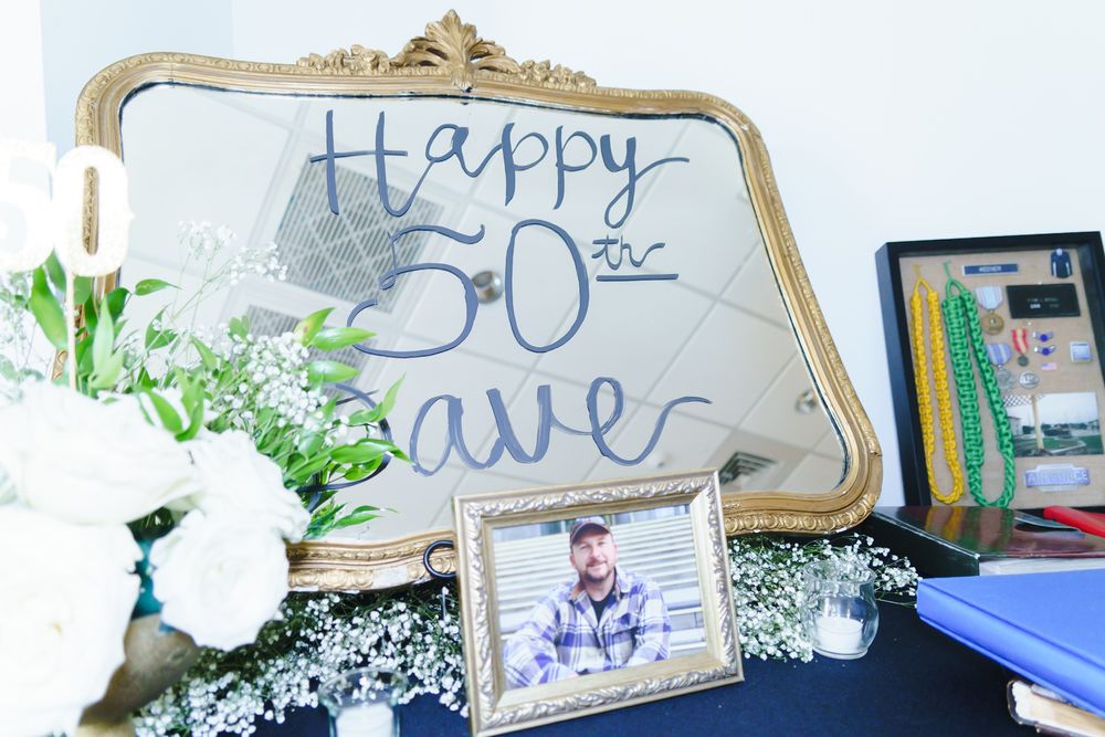 Gold framed mirror with Happy 50th Dave written in blue, beside white flowers and a framed photo at a birthday party.