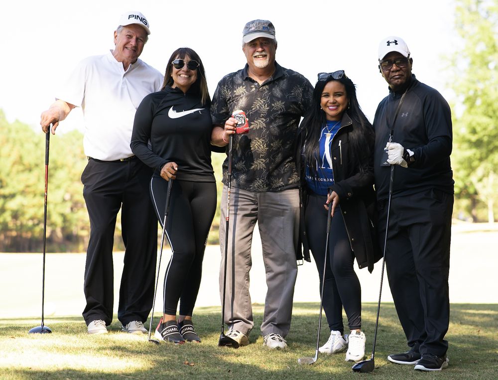 Five people posing together on a golf course, smiling for a group photo on a sunny day.
