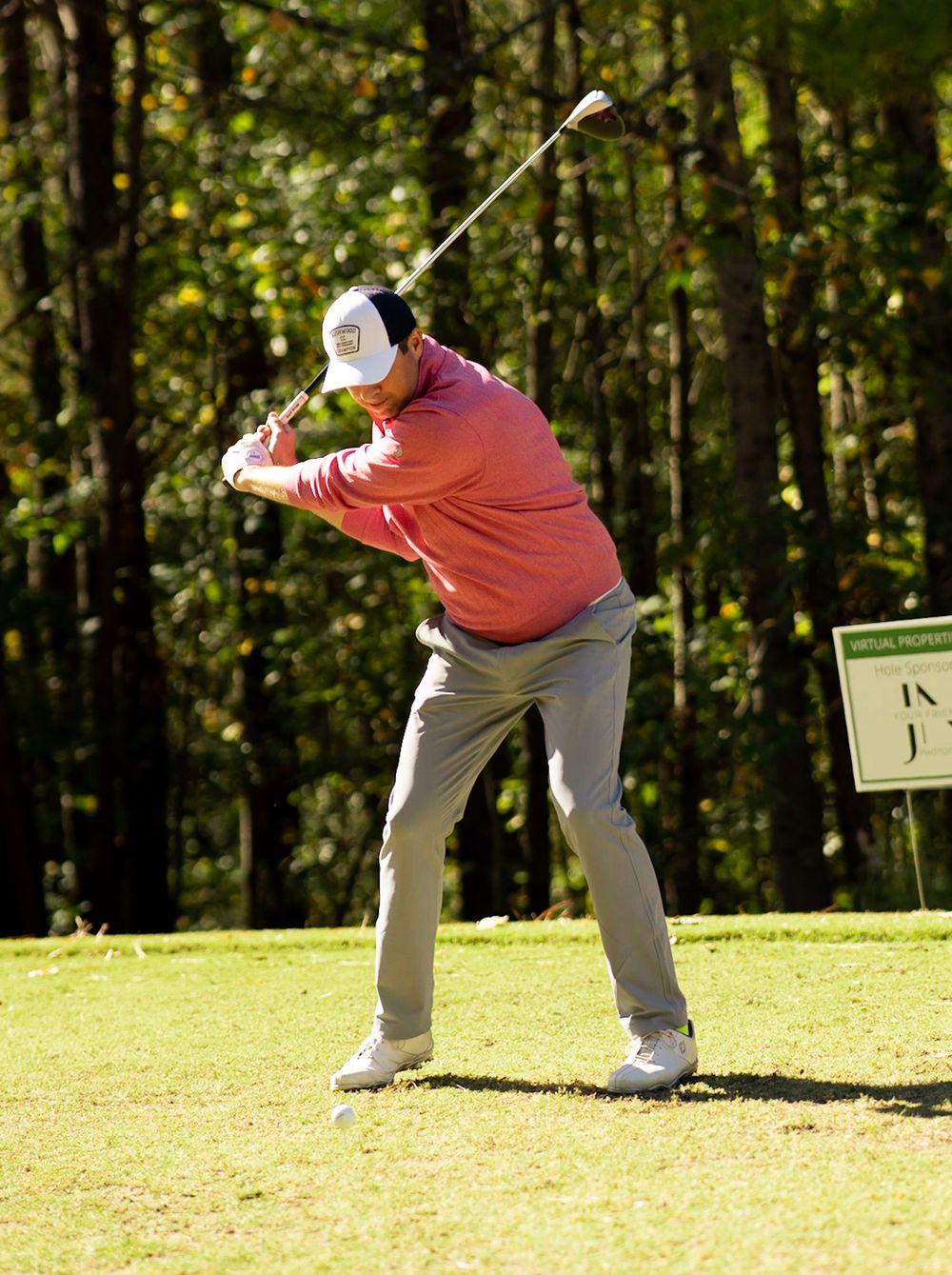 Golfer in pink sweater and white cap completing a full swing on a golf course surrounded by tall trees.
