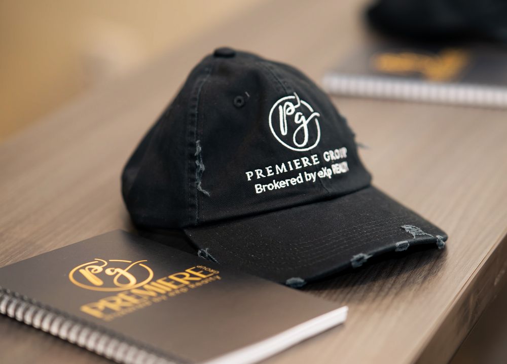Black distressed Premiere Group brokered by eXp Realty cap resting on a branded notebook on a desk.