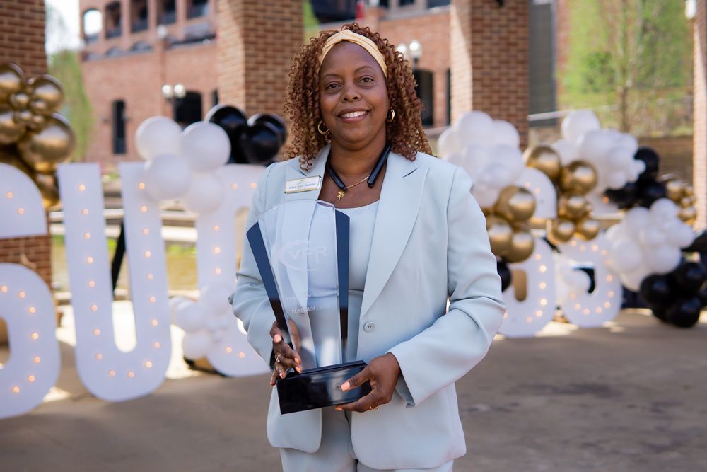 Woman in light gray blazer holds trophy award outdoors near balloon arch with black and gold decorations.