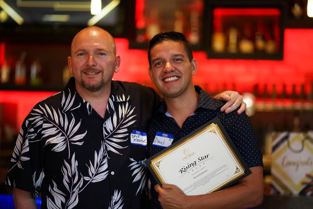 Two men smiling and posing together at a bar, one holding a framed certificate at an award recognition event.