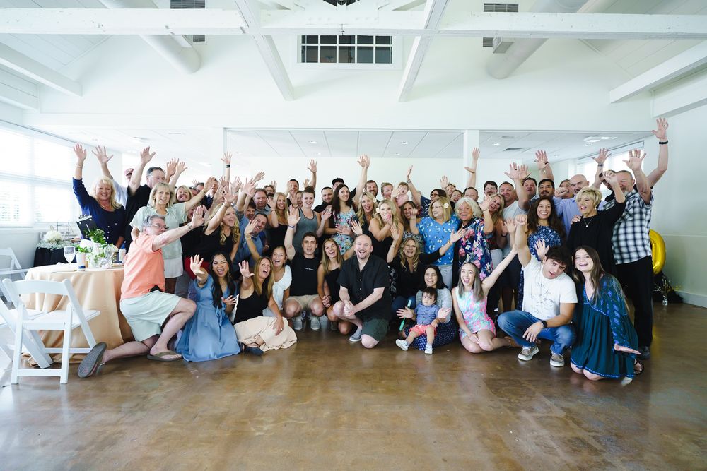 Large group of people smiling and raising hands at an indoor celebration event in a bright white venue.
