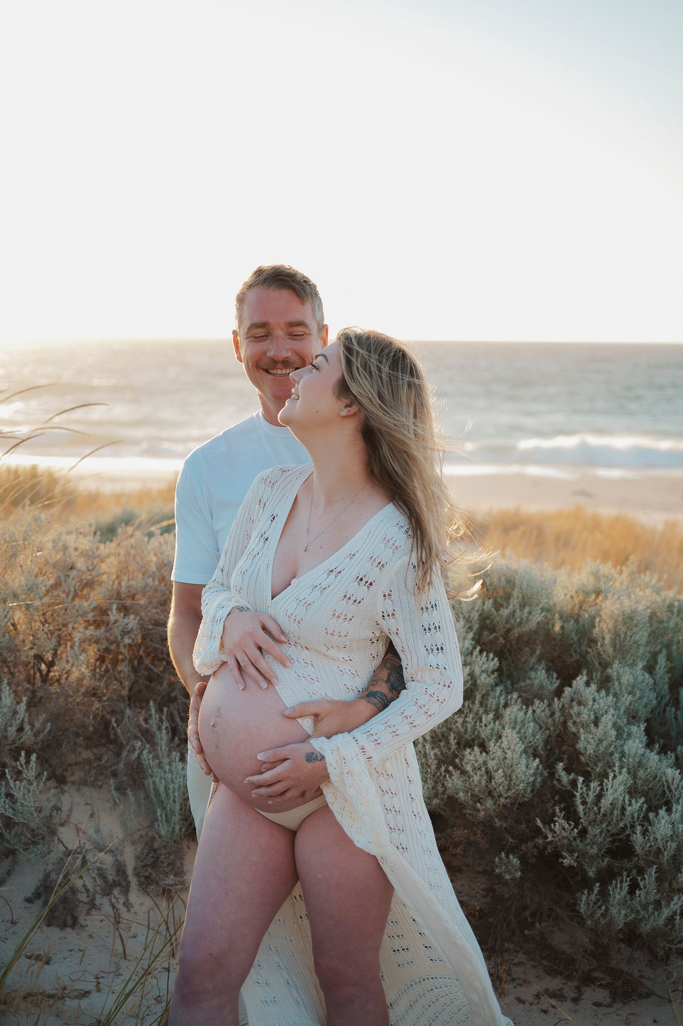 Couple embracing outdoors near a sandy beach with lush greenery and golden sunset light in the background.