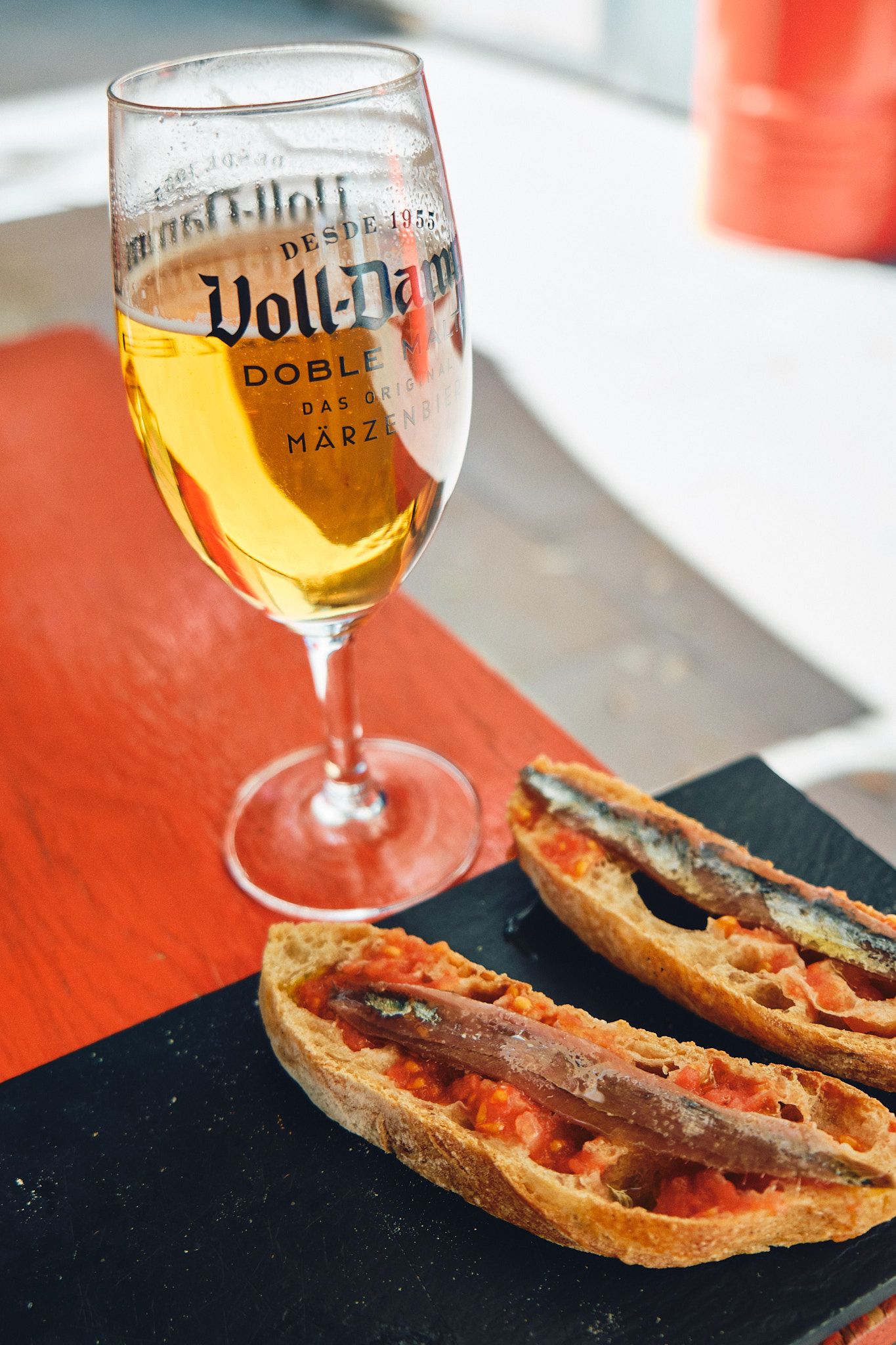 A glass of Voll-Damm beer beside two anchovy tomato toasts on a slate board.
