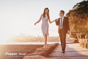 Couple walking hand-in-hand along a waterfront pier at sunset with city skyline in background.
