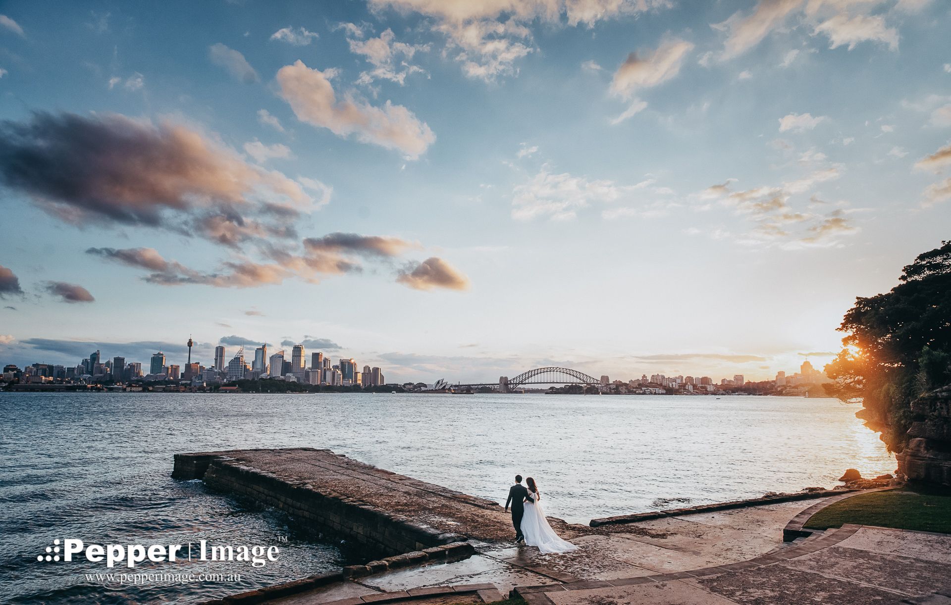 Couple poses on Bradleys Head Amphitheatre at sunset with Sydney skyline and Opera House in background.