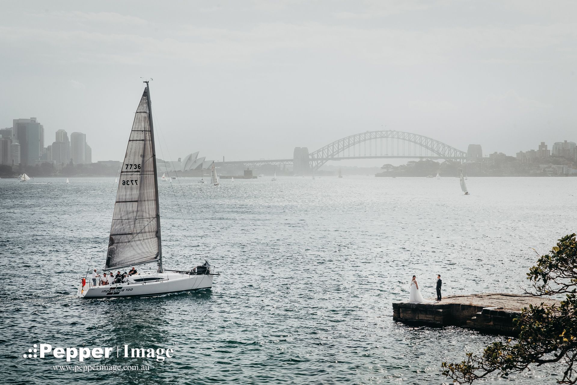 Sailboat on Sydney Harbour with Opera House and Harbour Bridge in misty background, couple standing on Bradleys Head Amphitheatre.