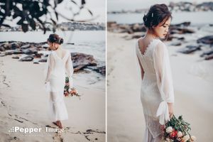 Bride in white lace long-sleeve gown holding bouquet on sandy beach with rocks and ocean in background.