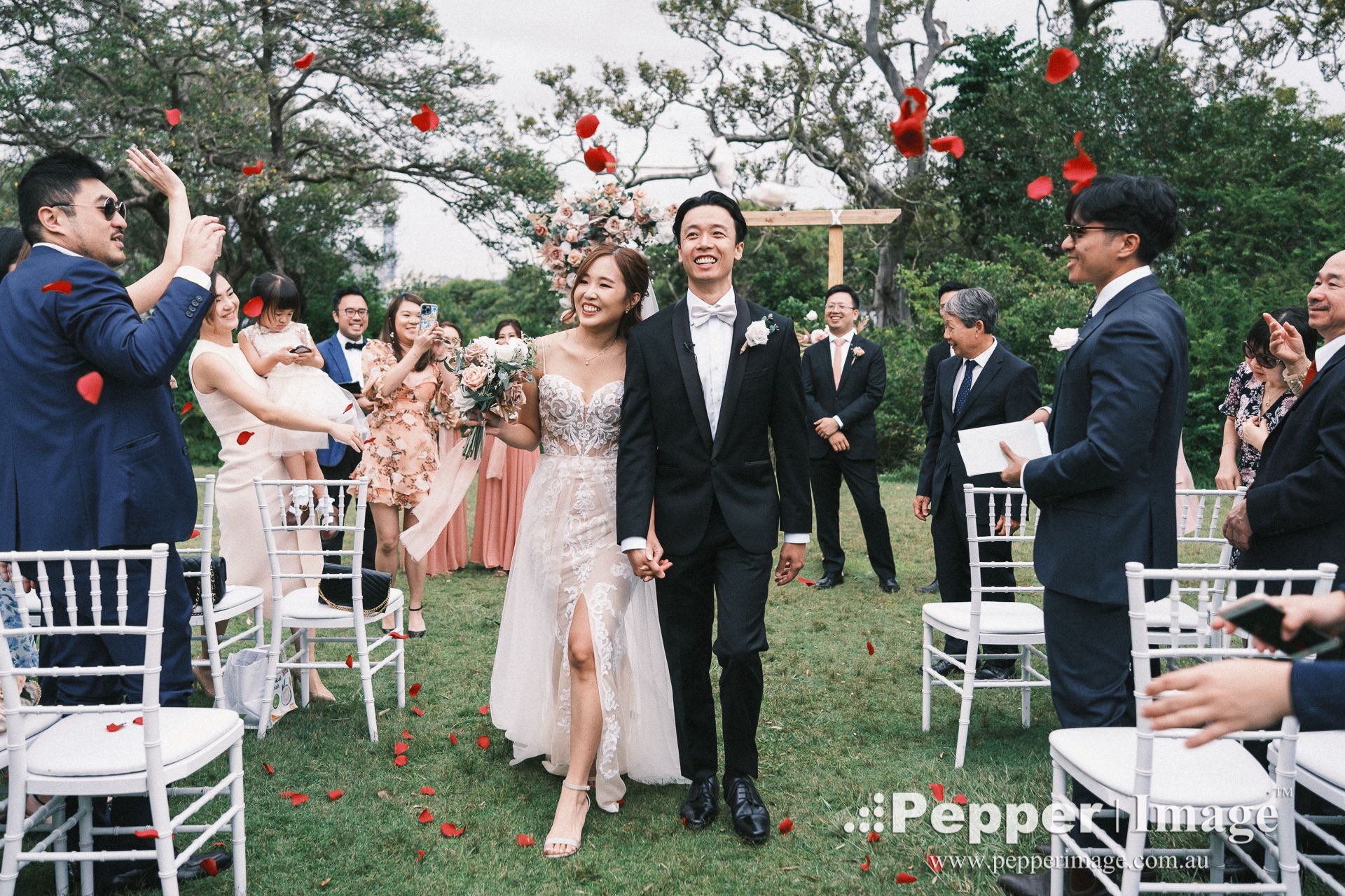 Couple walks down outdoor wedding aisle as guests toss rose petals, bride in pink gown holding bouquet.