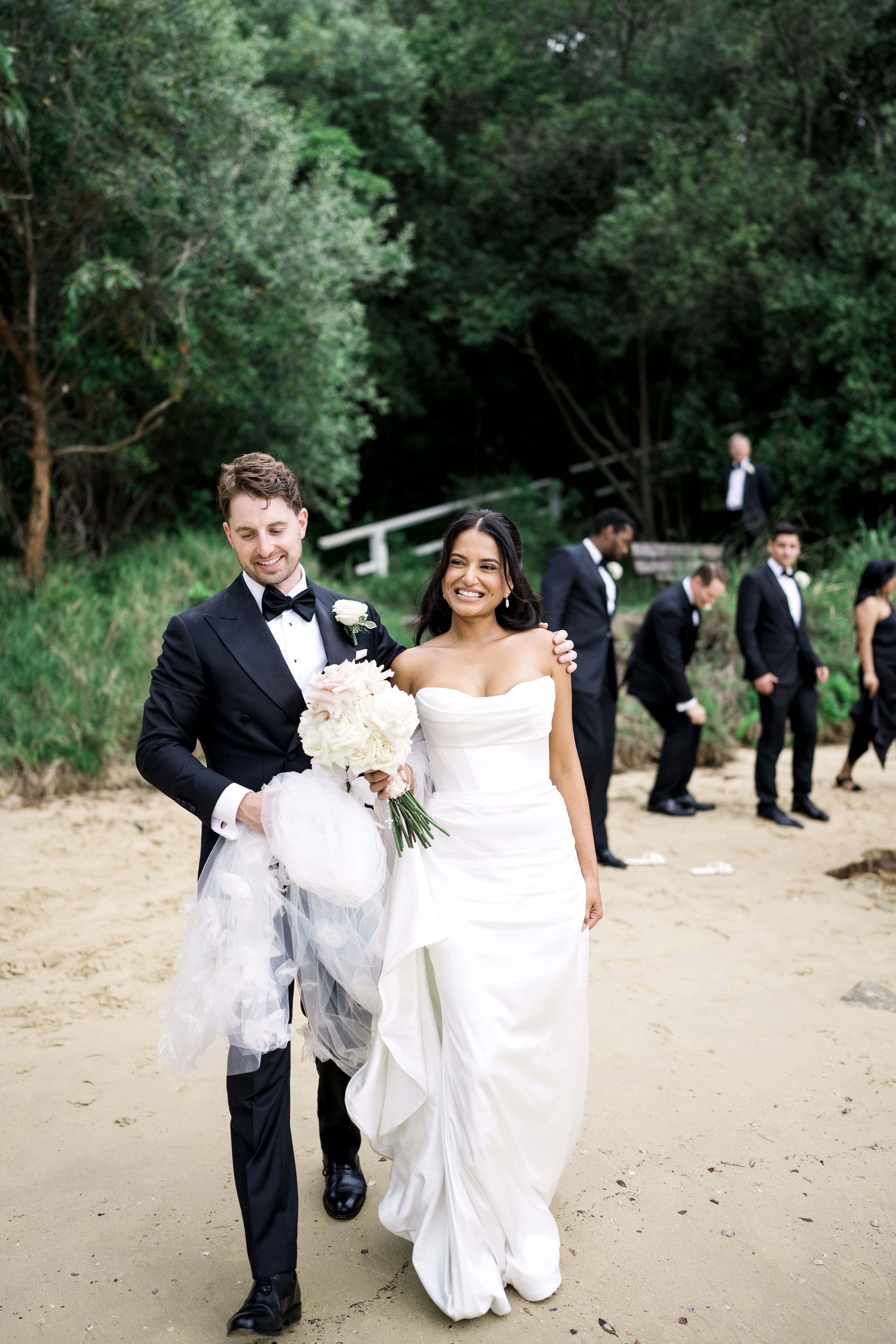 Bride in strapless white gown and groom in black tuxedo walk on Athol beach, holding bouquet, with groomsmen in background.