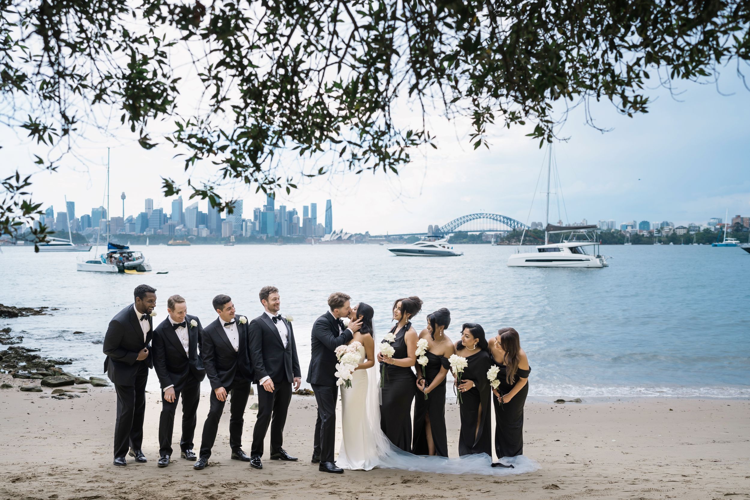 Wedding party poses on Sydney Harbour beach with Opera House and city skyline backdrop, bridal party in black and white attire.