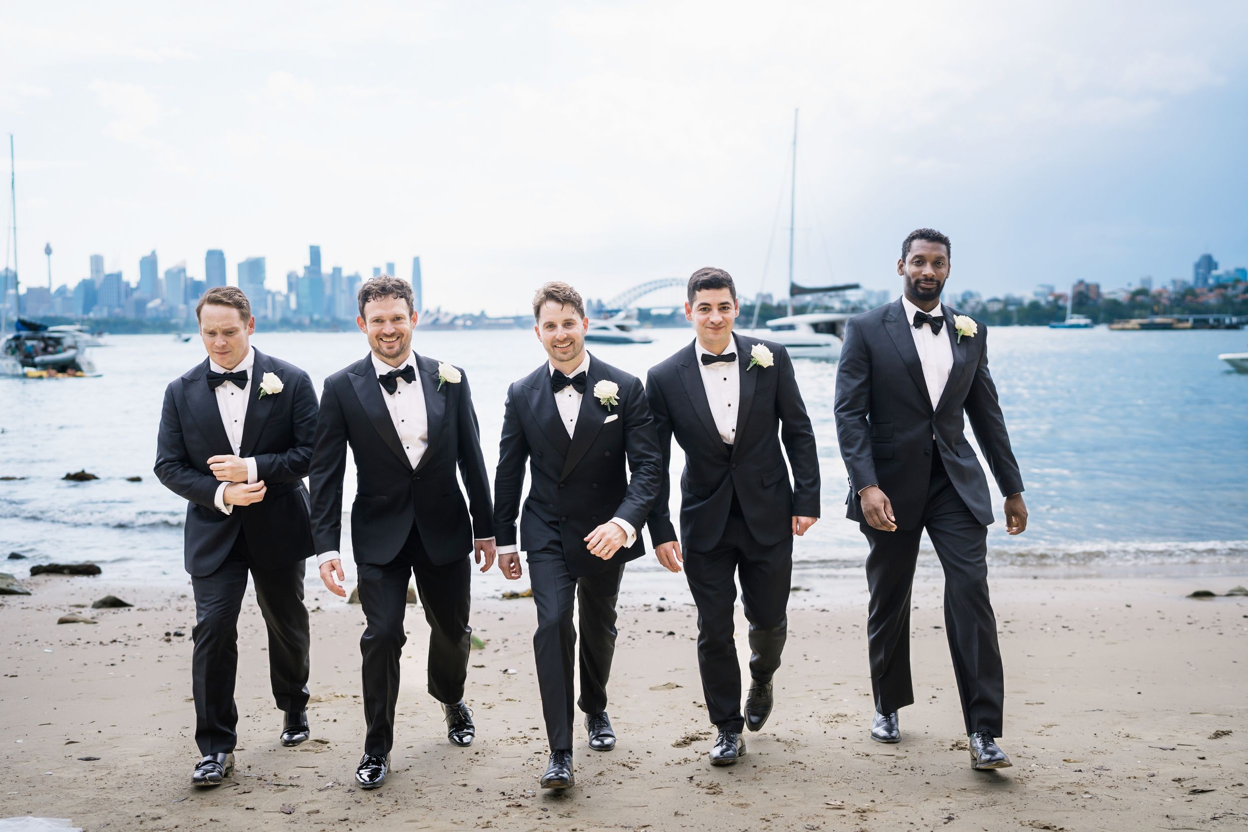 Five groomsmen in black tuxedos with bow ties walk along Athol beach with Sydney skyline and harbor in the background.