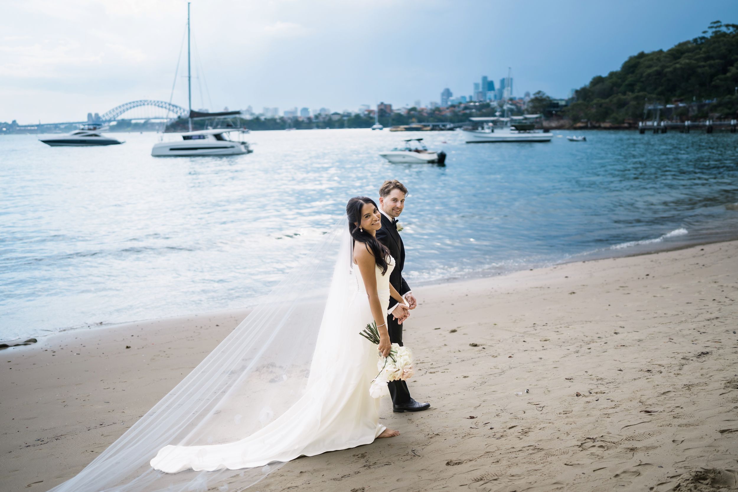 Bride and groom posing on Athol beach with Sydney Harbour Bridge and sailboats in background on wedding day.