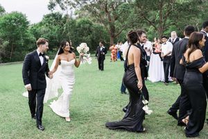 Bride and groom walk hand-in-hand through cheering guests outdoors, bride in white gown holding bouquet, guests in black attire.