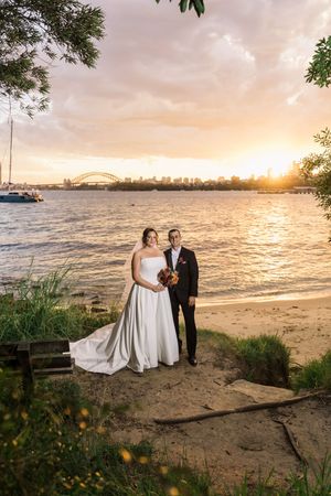Bride and groom pose on Athol beach at sunset with Harbour Bridge backdrop.