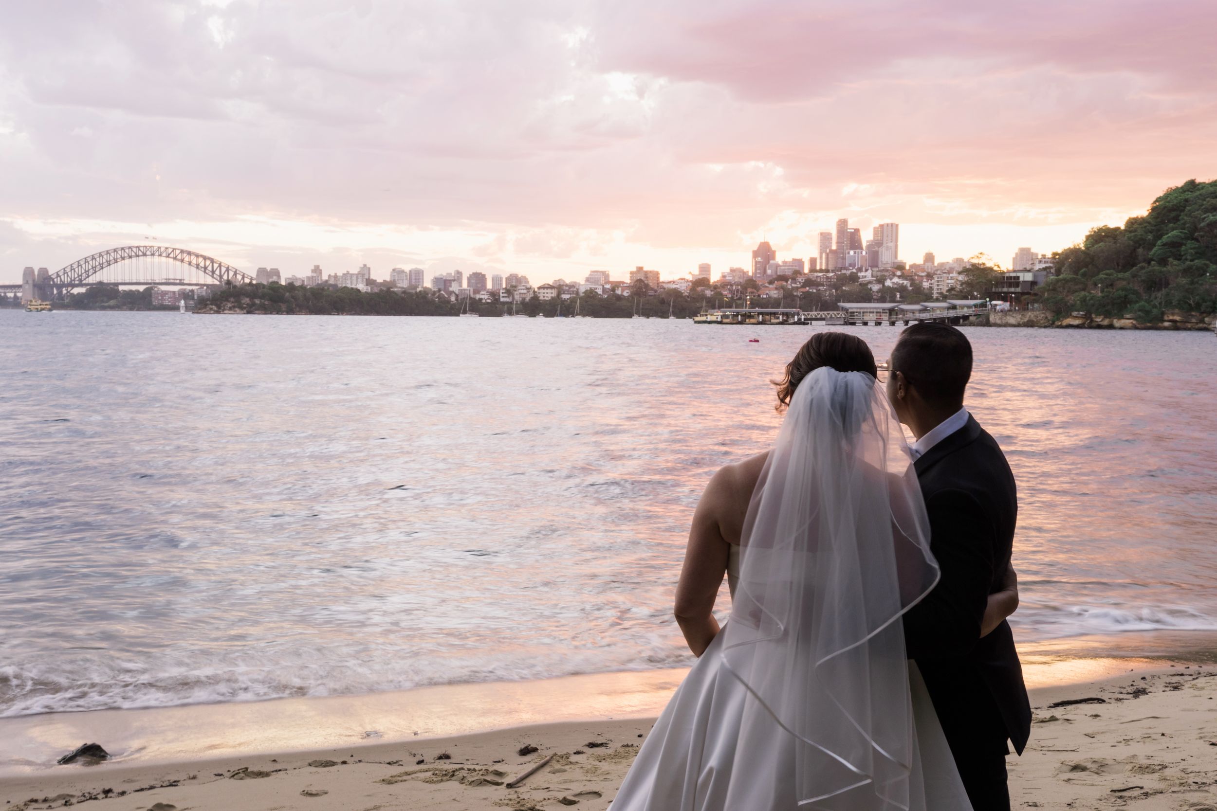Bride and groom stand on Athol Beach at sunset with Sydney Harbour Bridge and city skyline in the background.