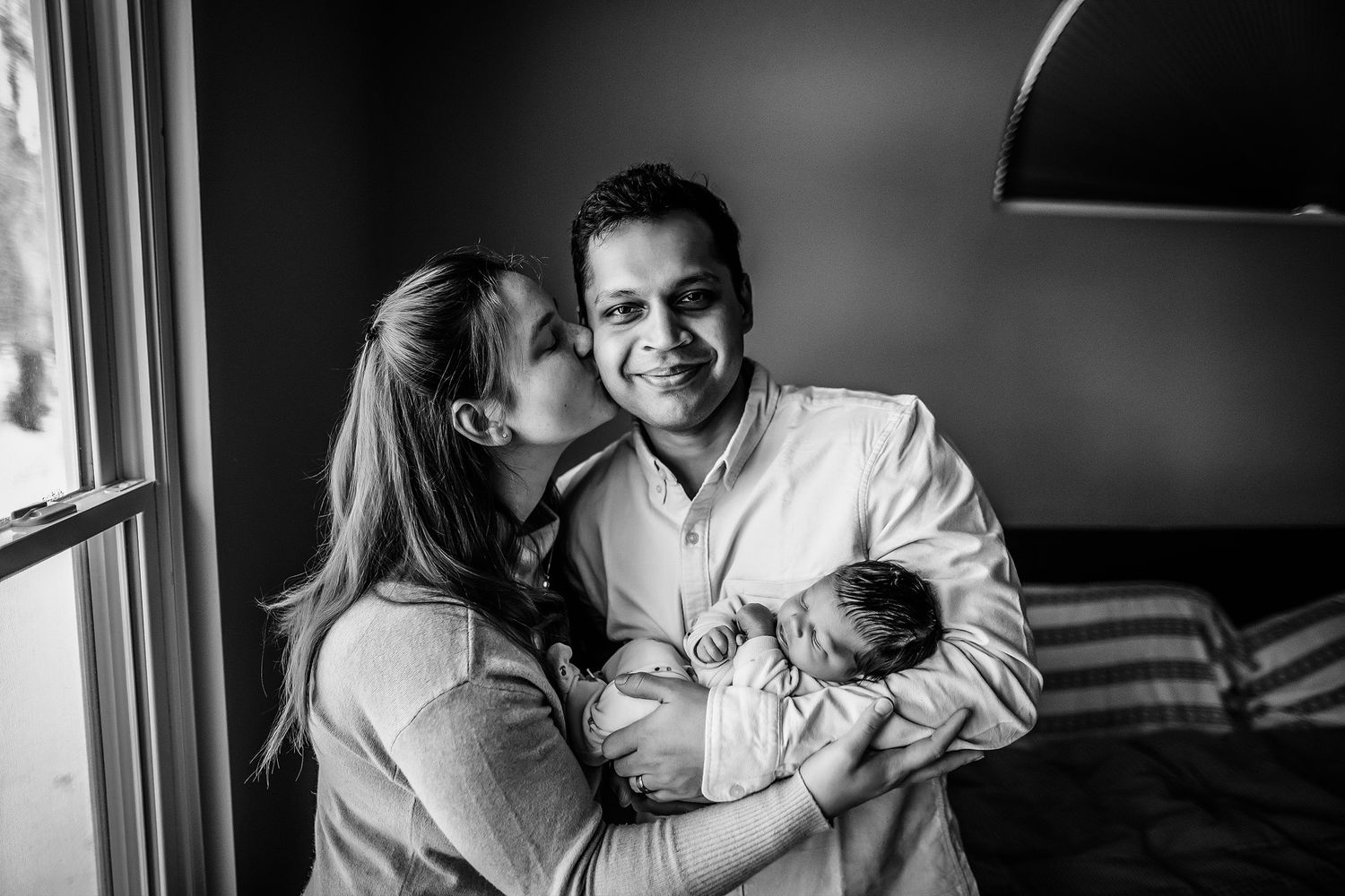 Black and white photo of a couple with their newborn baby, woman kissing man on cheek while he holds the infant.