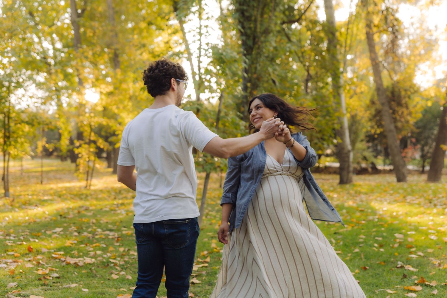 Couple dancing joyfully in a park with autumn trees, woman in striped dress and denim jacket holding hands with man.