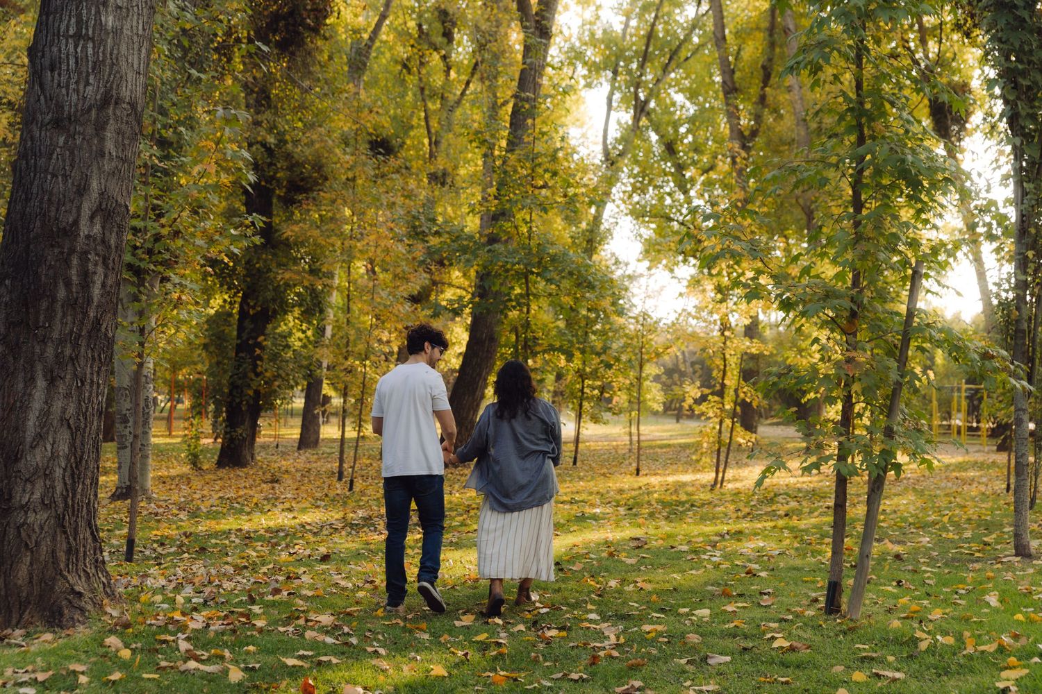 Couple holding hands walking through a scenic autumn park with yellow and green trees and fallen leaves on the ground.