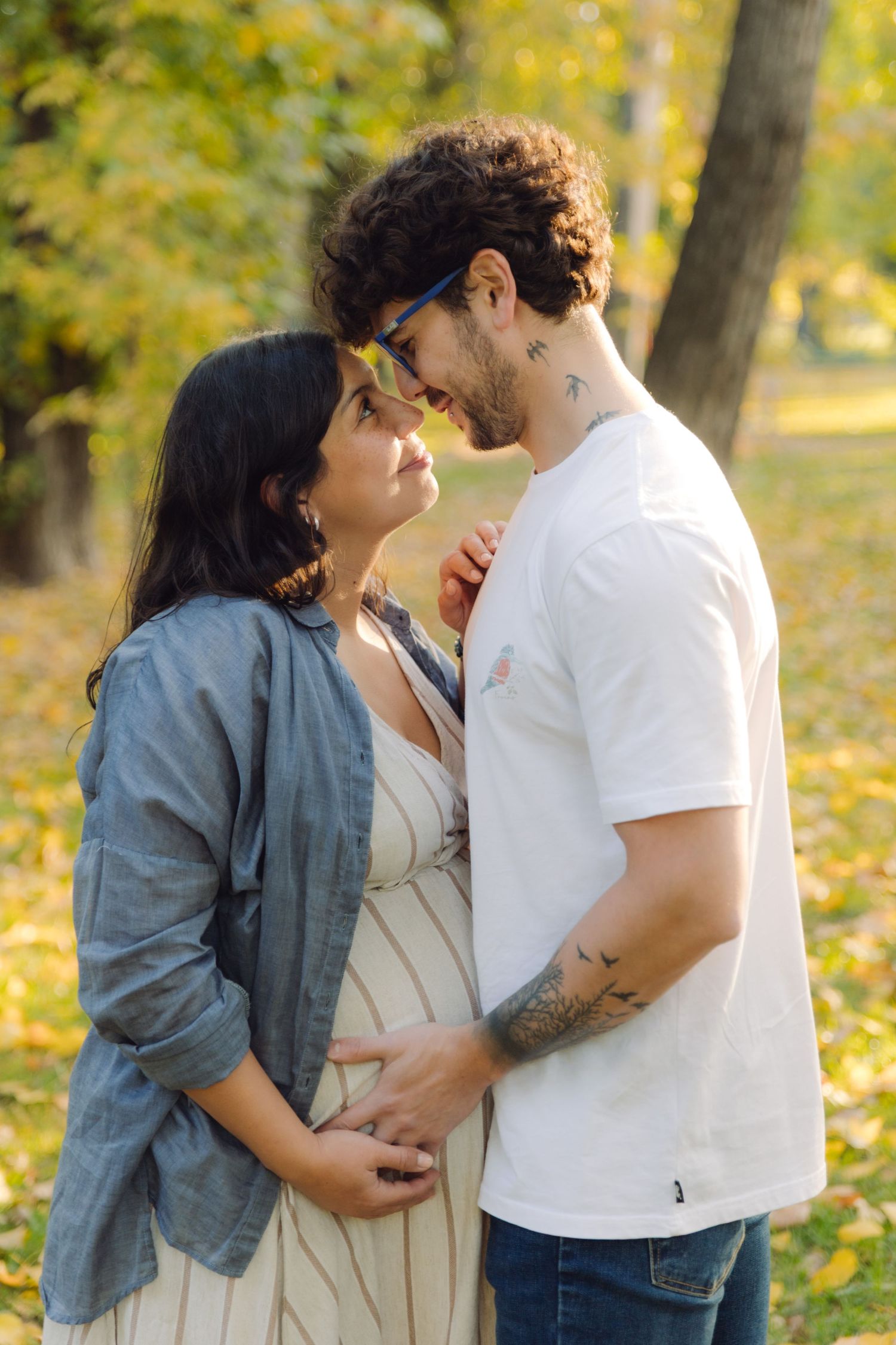 A couple shares a tender moment in an autumn park, the woman visibly pregnant as her partner gently holds her belly.