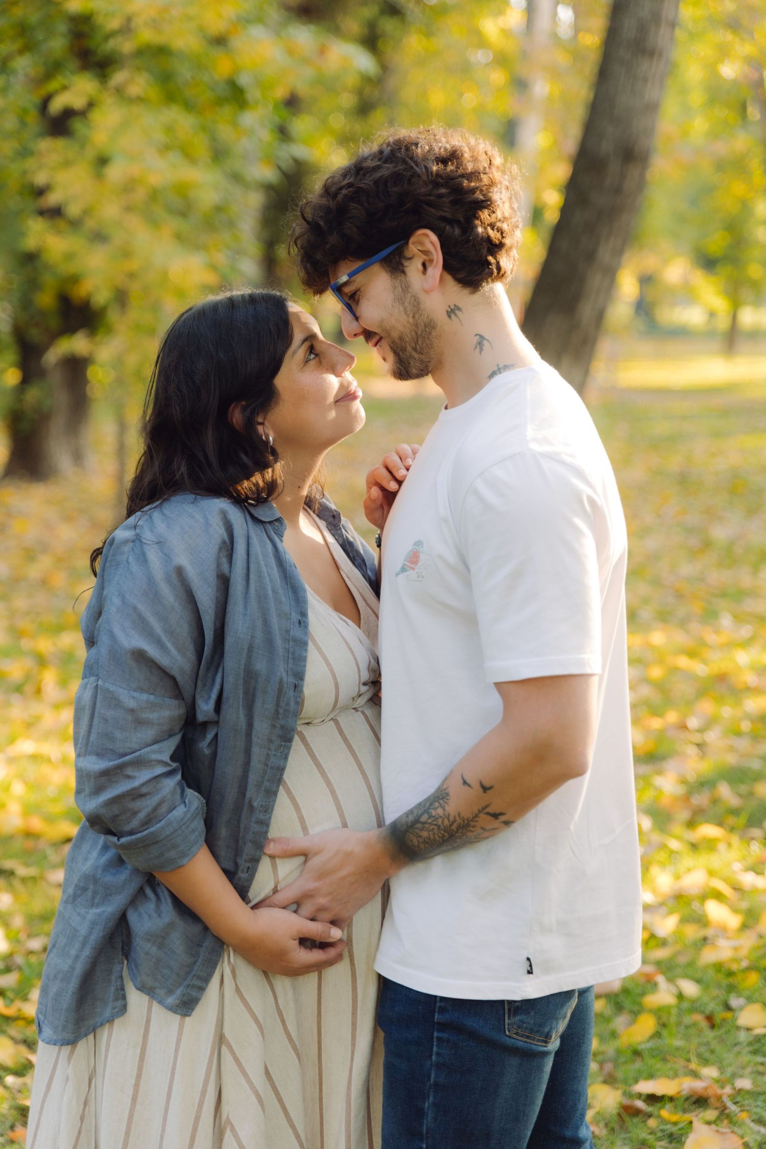 Pregnant couple embracing in autumn park, man with tattoos and curly hair smiling at woman in striped dress.