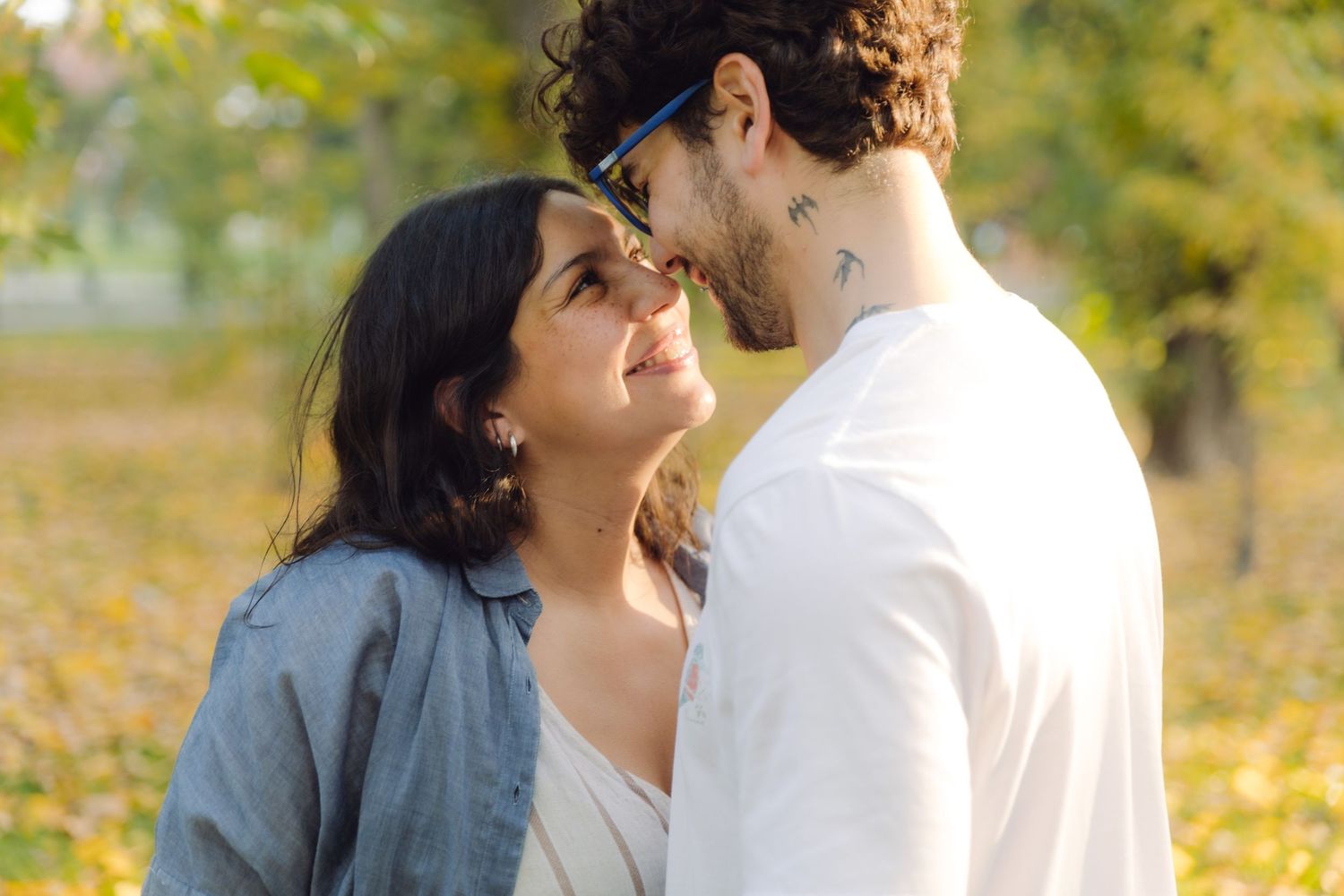A couple sharing a romantic moment outdoors in a park with autumn foliage in the background.
