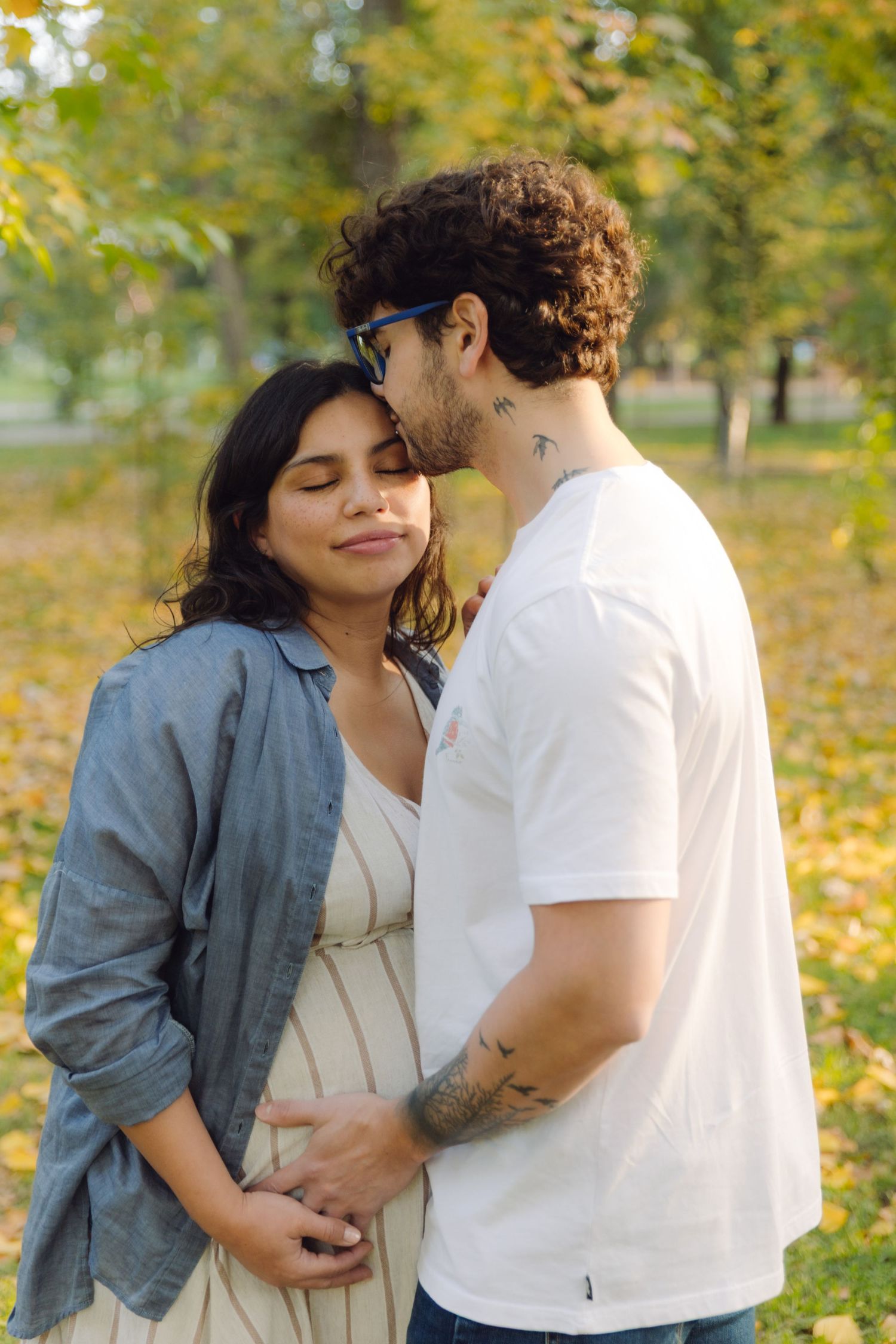 A couple embraces tenderly in an autumn park, the pregnant woman smiling as her partner kisses her forehead.