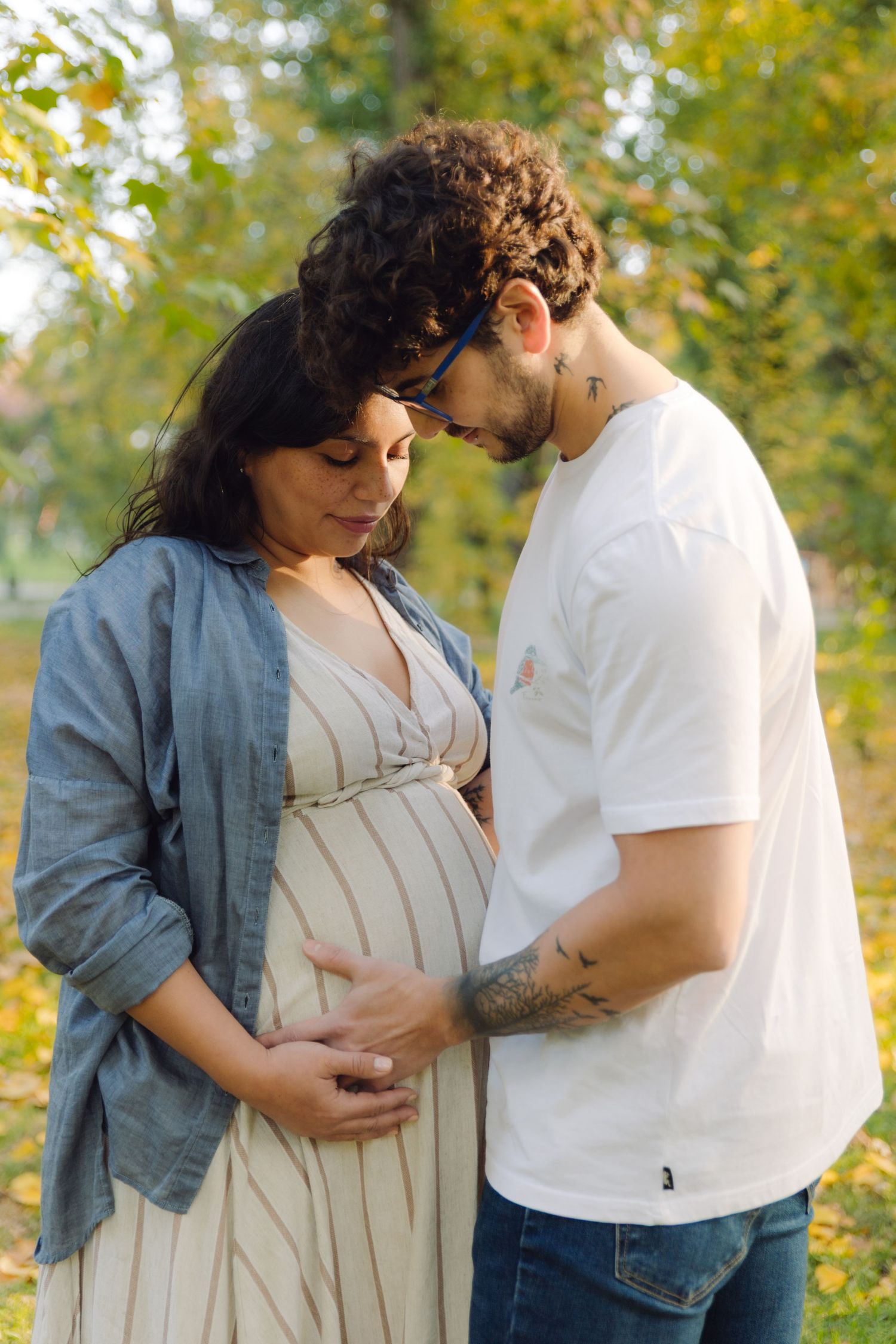 Expectant couple tenderly cradling pregnant belly in autumn park, man in white tee and woman in striped dress.