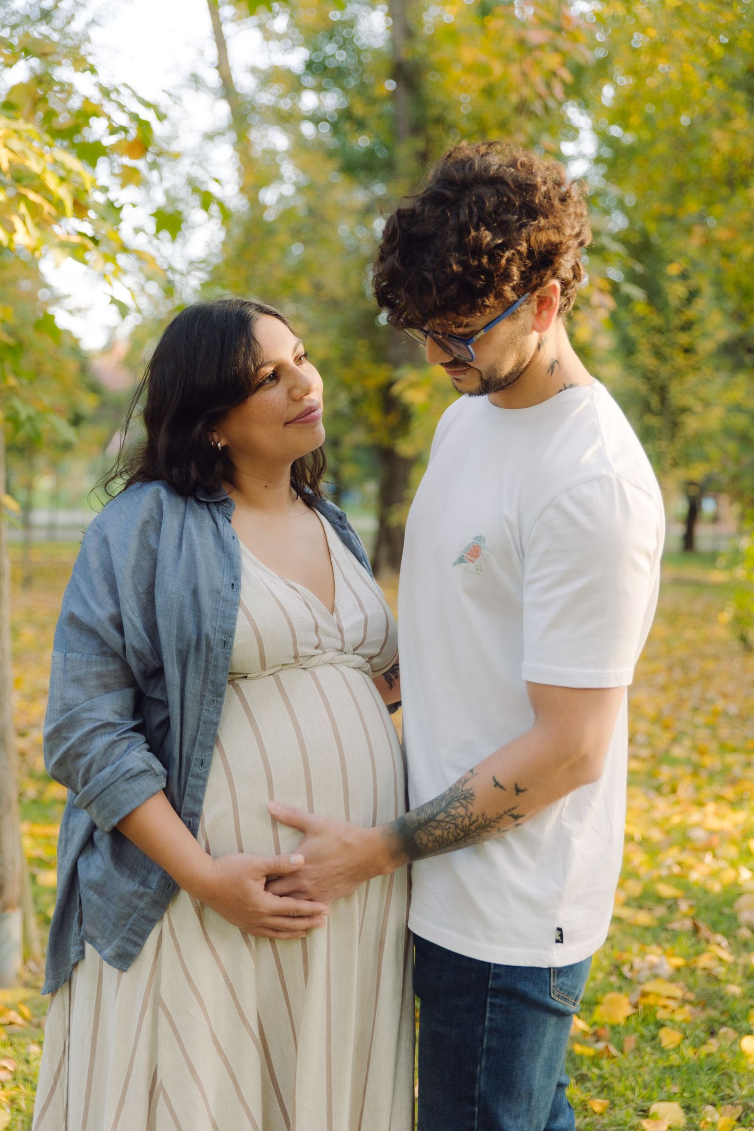 Couple in autumn park, pregnant woman in striped dress with partner in white tee gently cradling her baby bump.