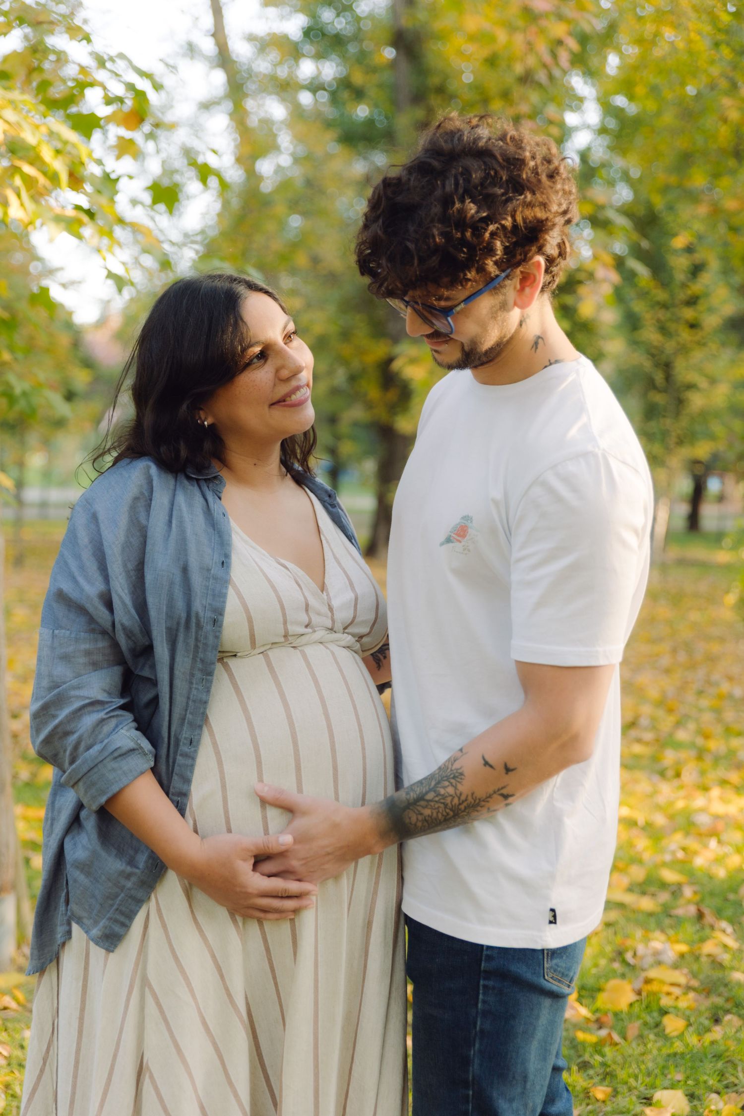 Pregnant woman in striped dress with partner touching her baby bump in autumn park.