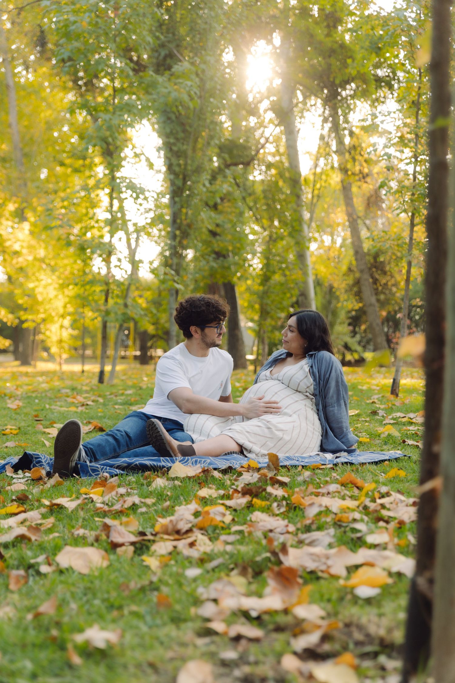 Couple sitting on blanket in autumn park, man touching pregnant woman's belly among fallen leaves in golden sunlight.