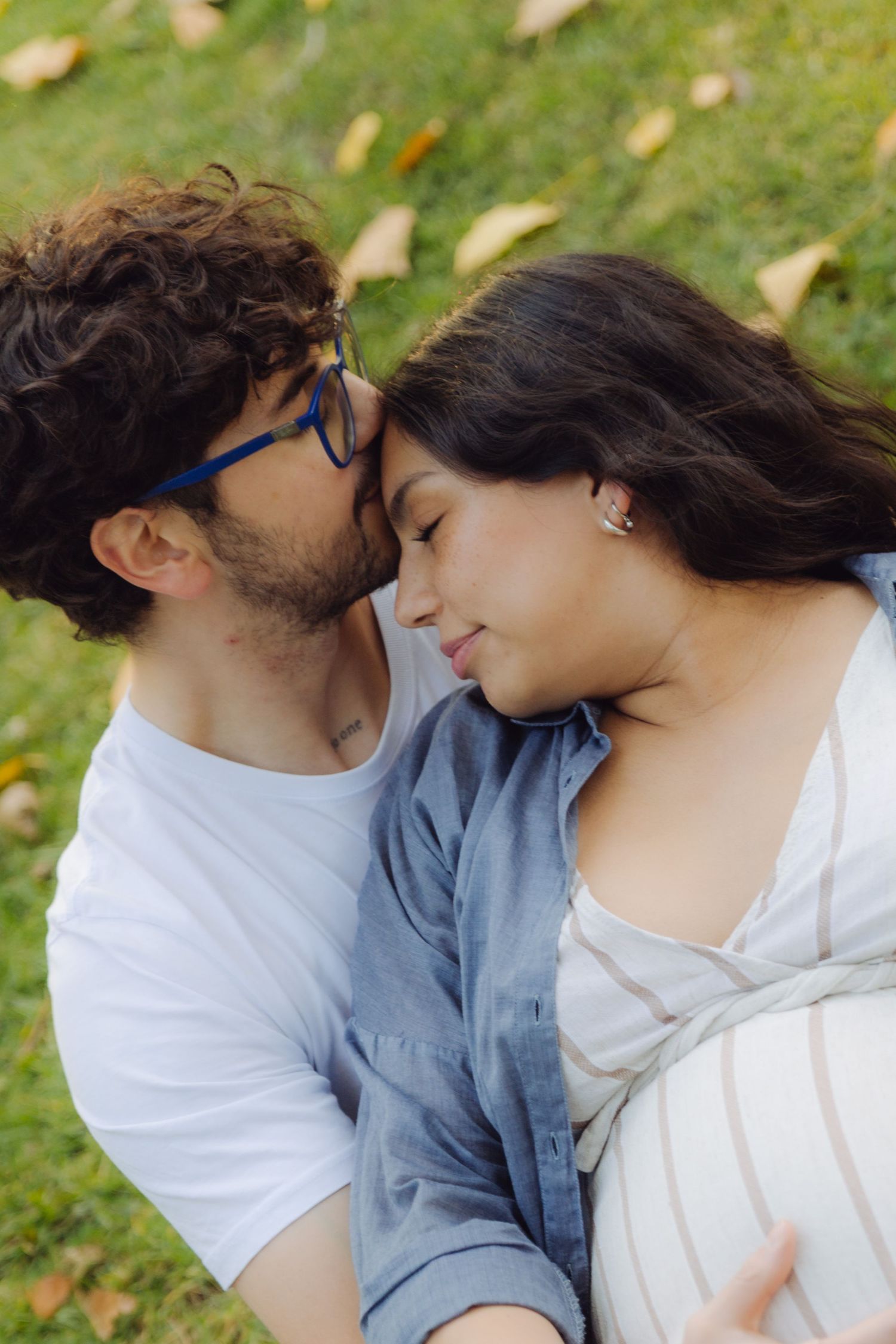 A couple shares a tender moment on grass, man with curly hair and blue glasses kissing woman's forehead.
