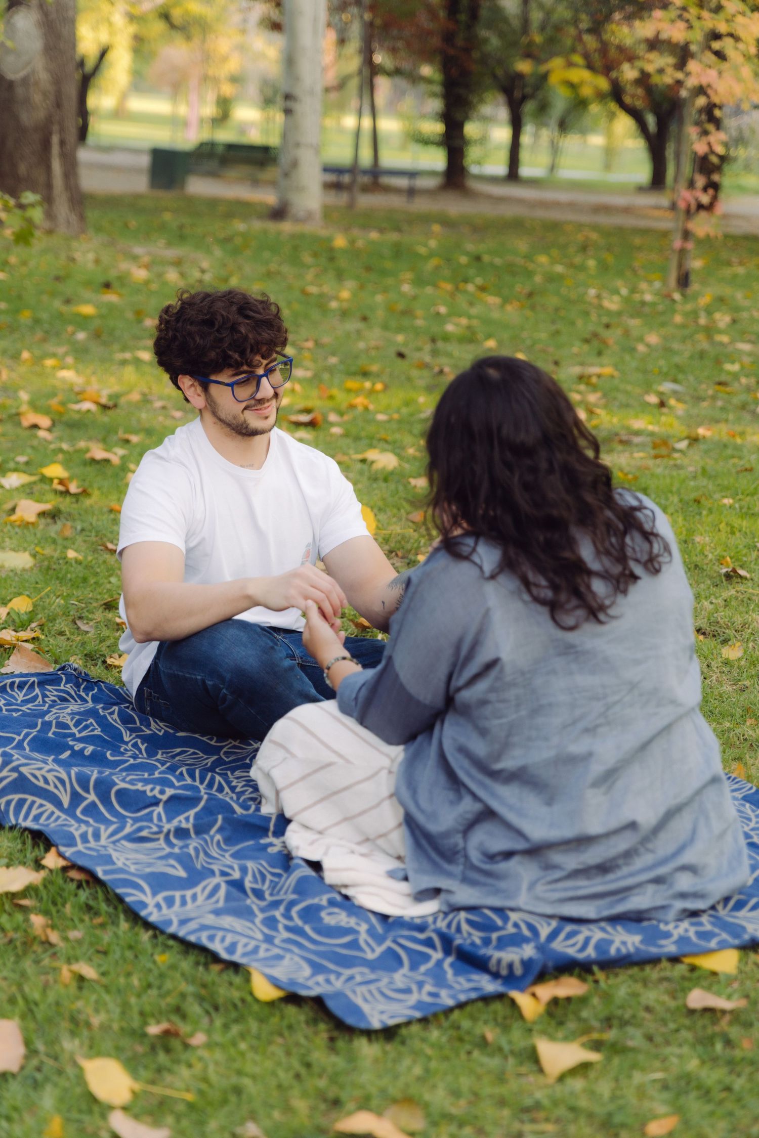Couple sitting on blue blanket in autumn park holding hands, surrounded by fallen yellow leaves on green grass.