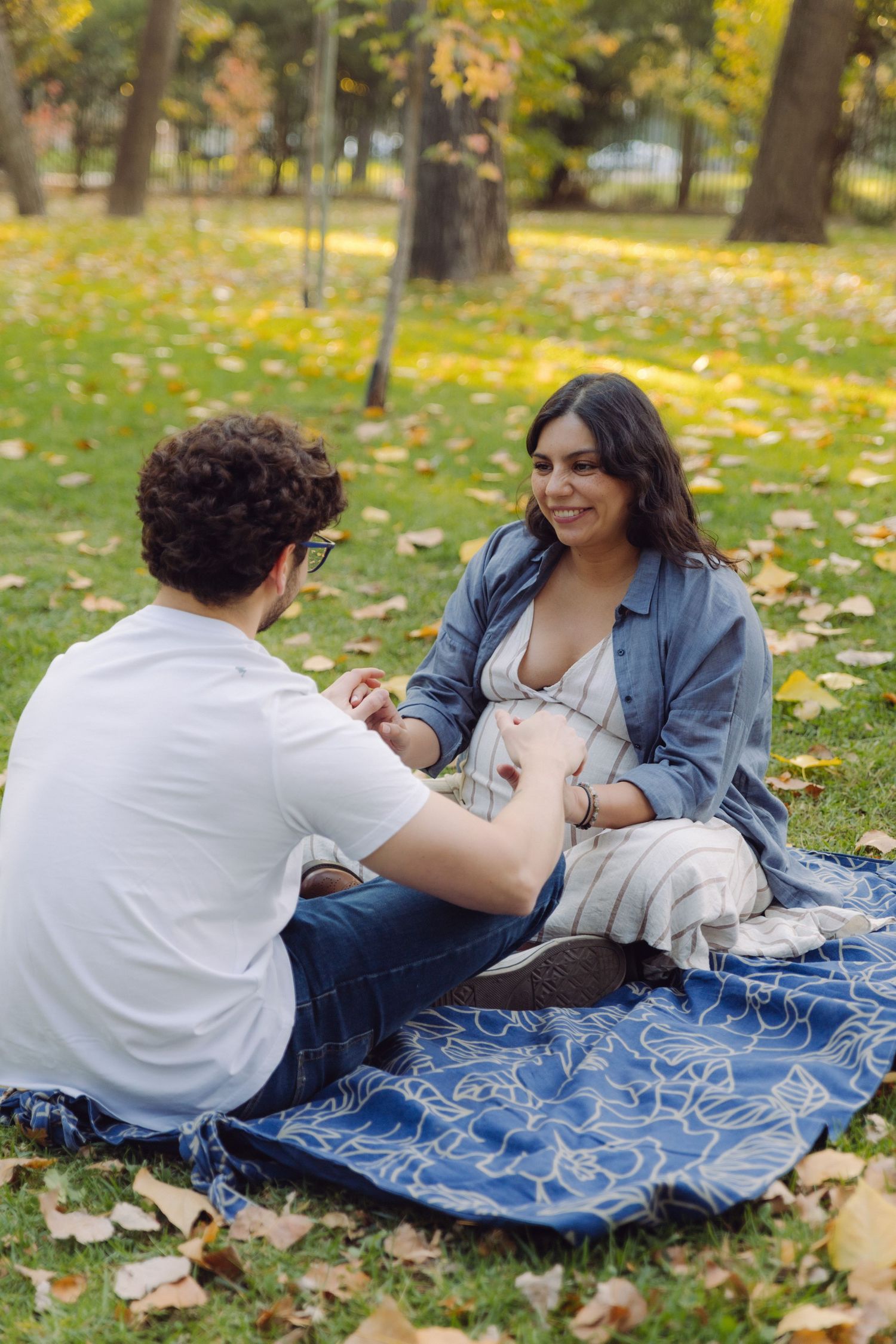 Pregnant woman smiling at partner while sitting on blanket in autumn park with fallen leaves.