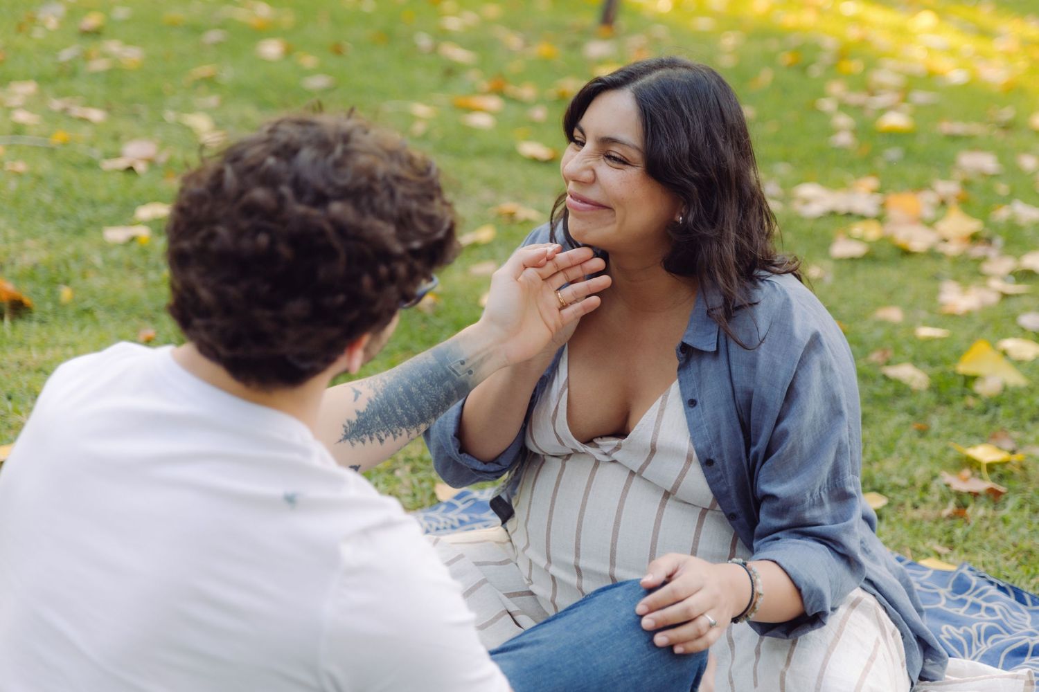 Pregnant woman in blue shirt sitting on grass in park, smiling as partner gently touches her face.