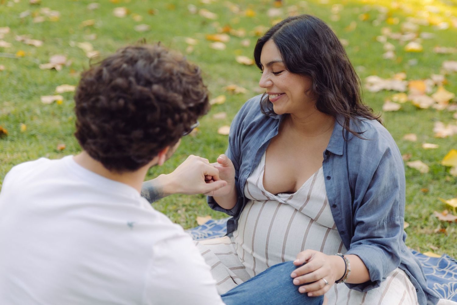 Pregnant woman sitting on grass in park sharing a tender moment with partner on sunny day.