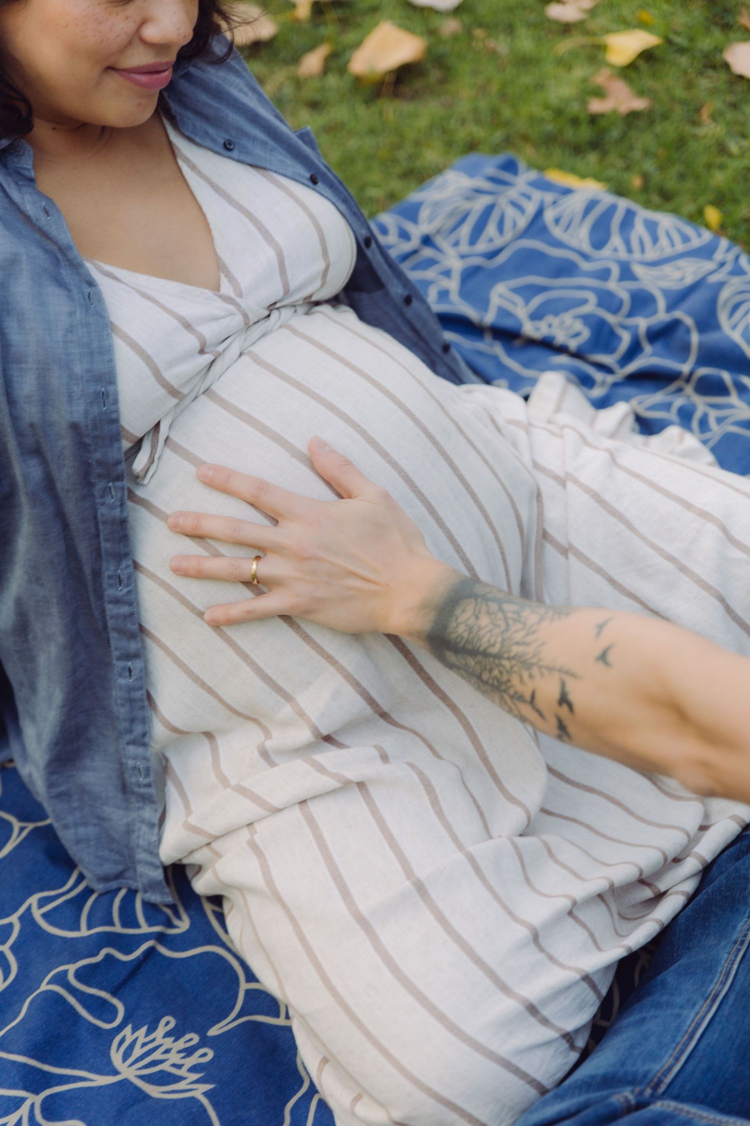 Pregnant woman in striped dress and denim jacket sitting on blue blanket outdoors, gently cradling her baby bump.