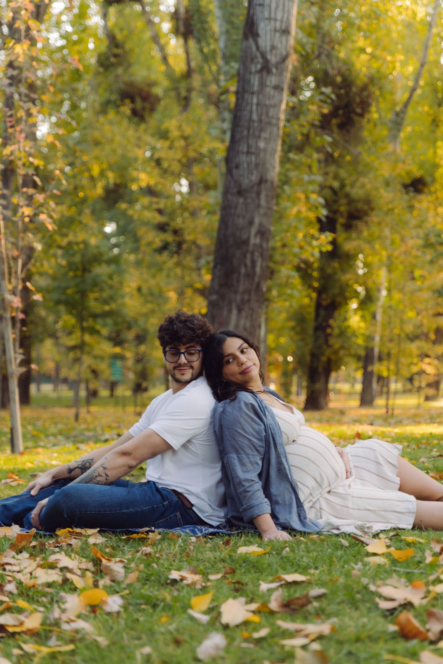 Couple sitting back-to-back on grass in autumn park, surrounded by fallen leaves and colorful trees.