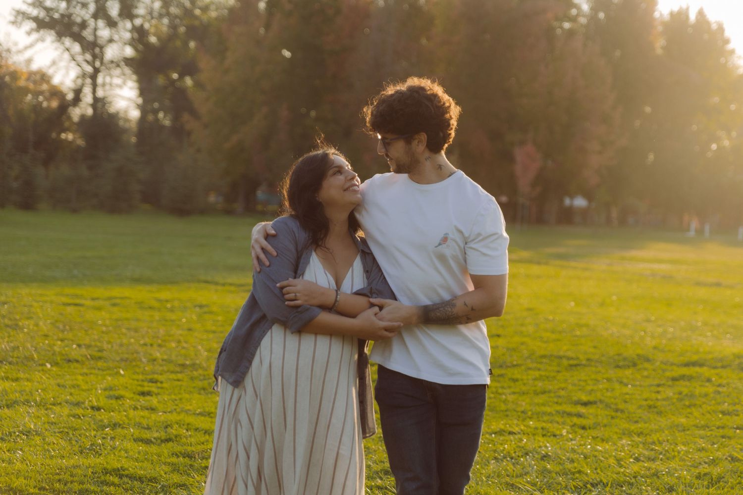 Couple embracing and smiling at each other in a sunny green park during golden hour.