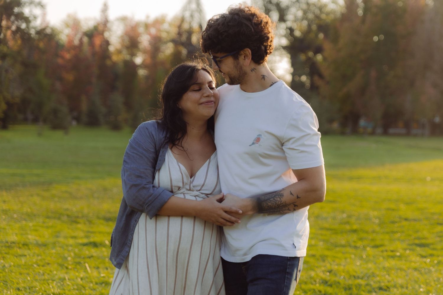 Couple embracing in a sunny park, pregnant woman in striped dress leaning on partner in white t-shirt.