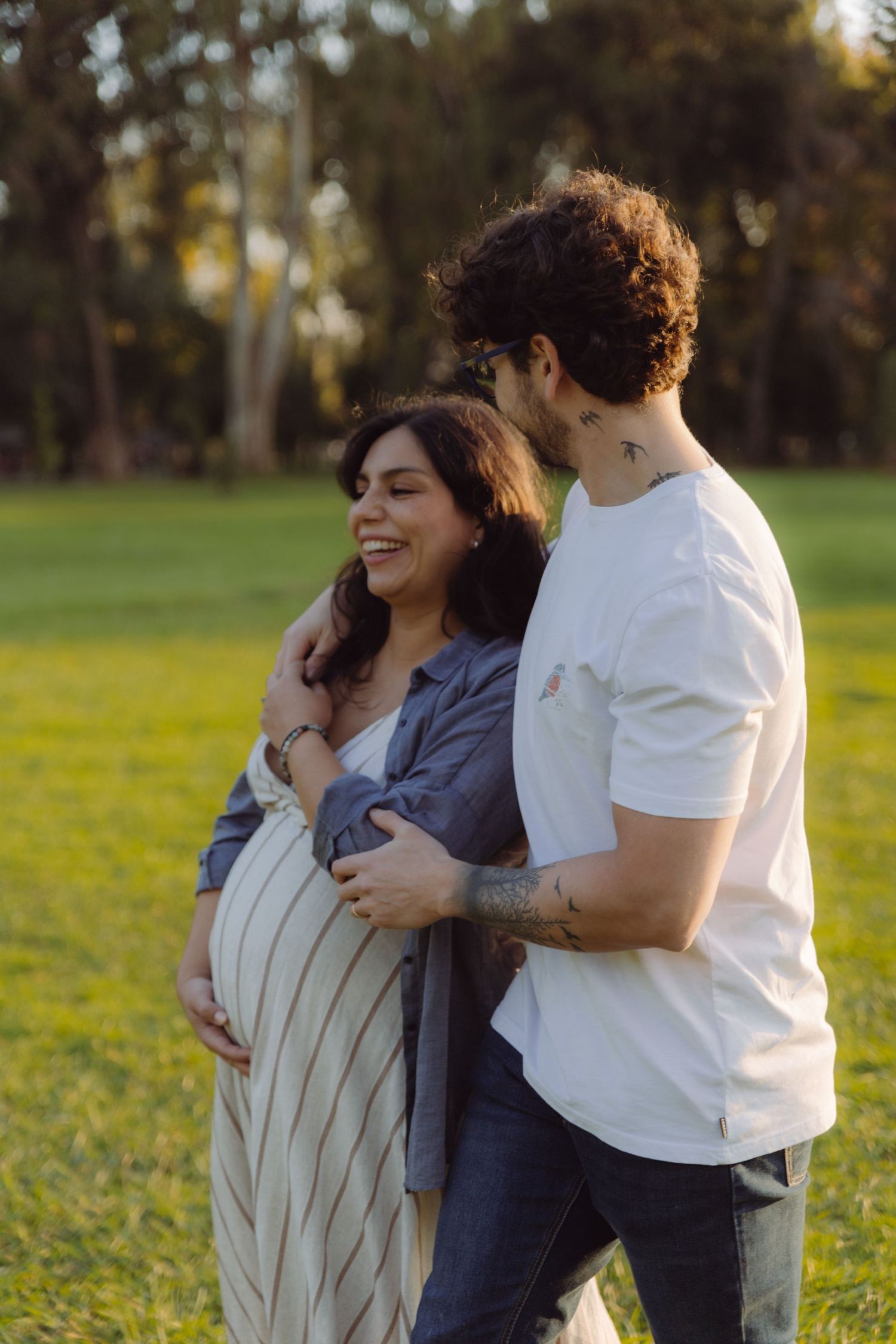 Smiling pregnant woman in striped dress embraced by curly-haired man in white tee in sunny park.