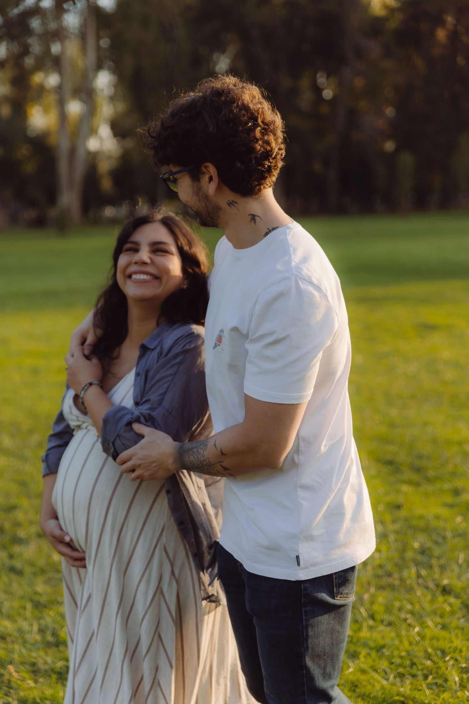 Couple embracing outdoors at golden hour, pregnant woman smiling as man hugs her belly in a green park.