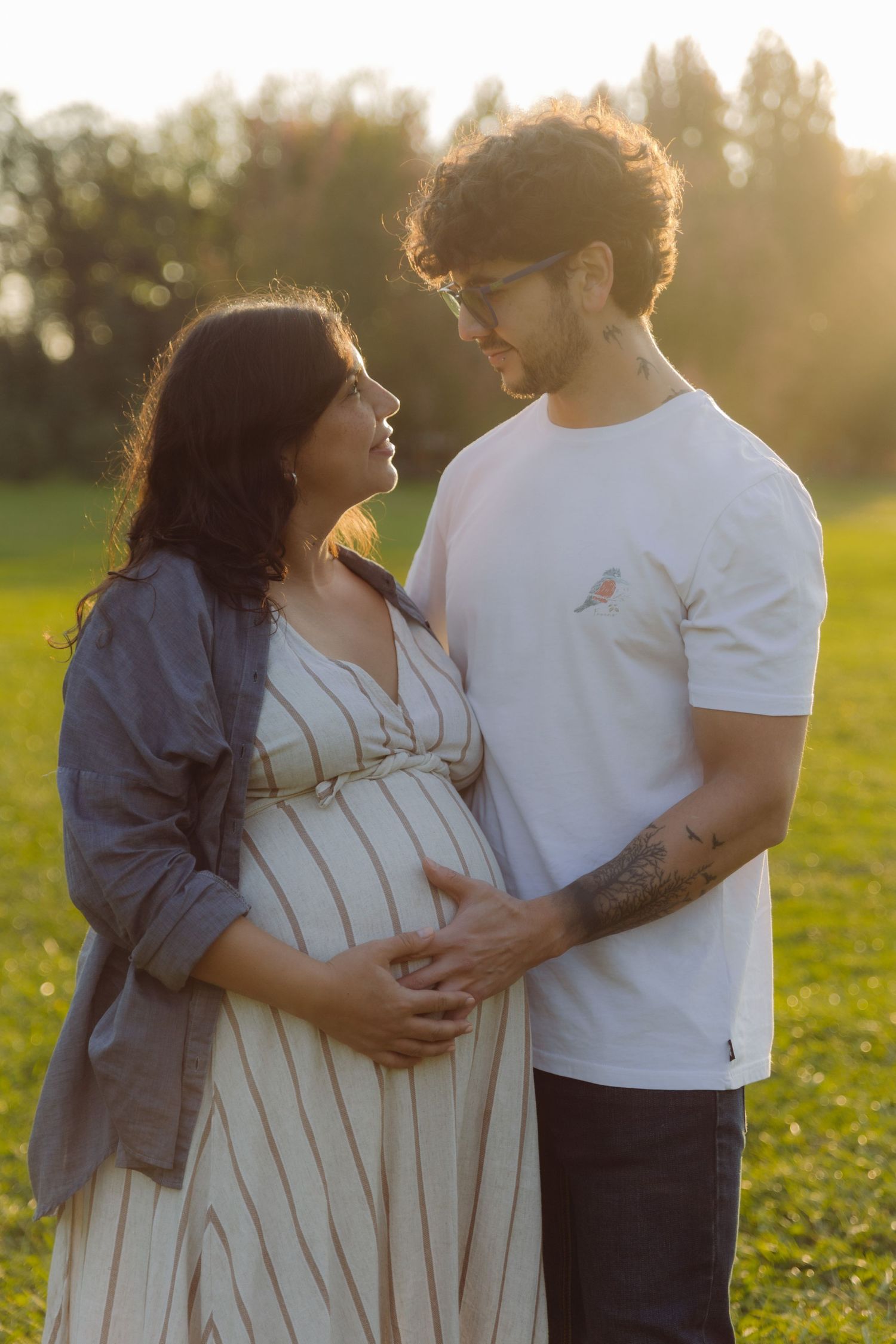 Expecting couple standing in sunlit park, man in white tee embracing pregnant woman in striped dress at golden hour.