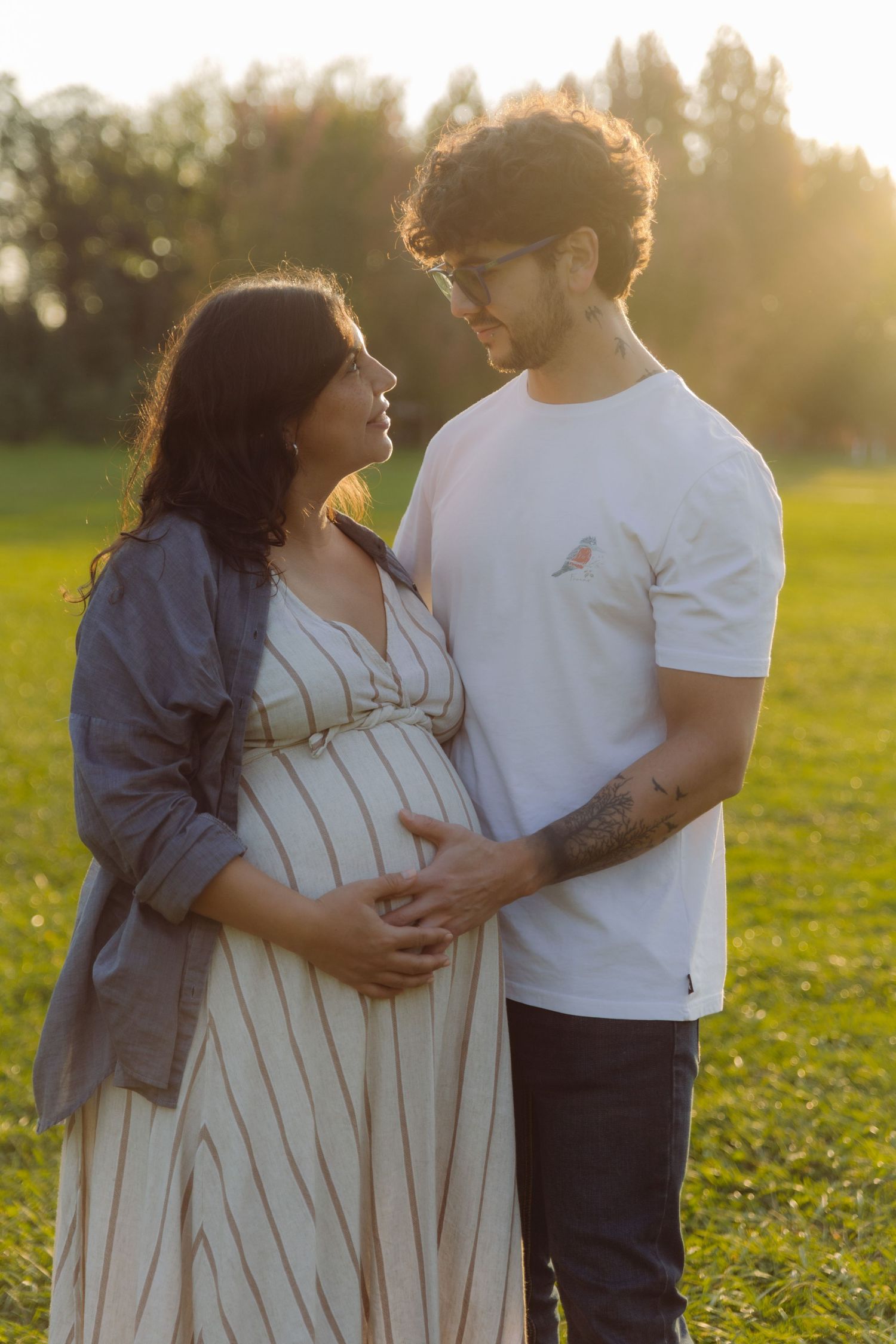 Couple embracing at sunset in green field, man holding pregnant woman's belly tenderly.