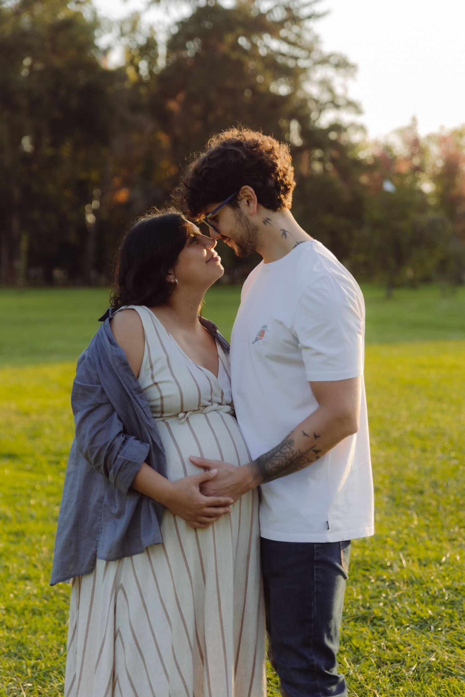 Expecting couple sharing tender moment, facing each other in golden sunlit park, man cradling baby bump.