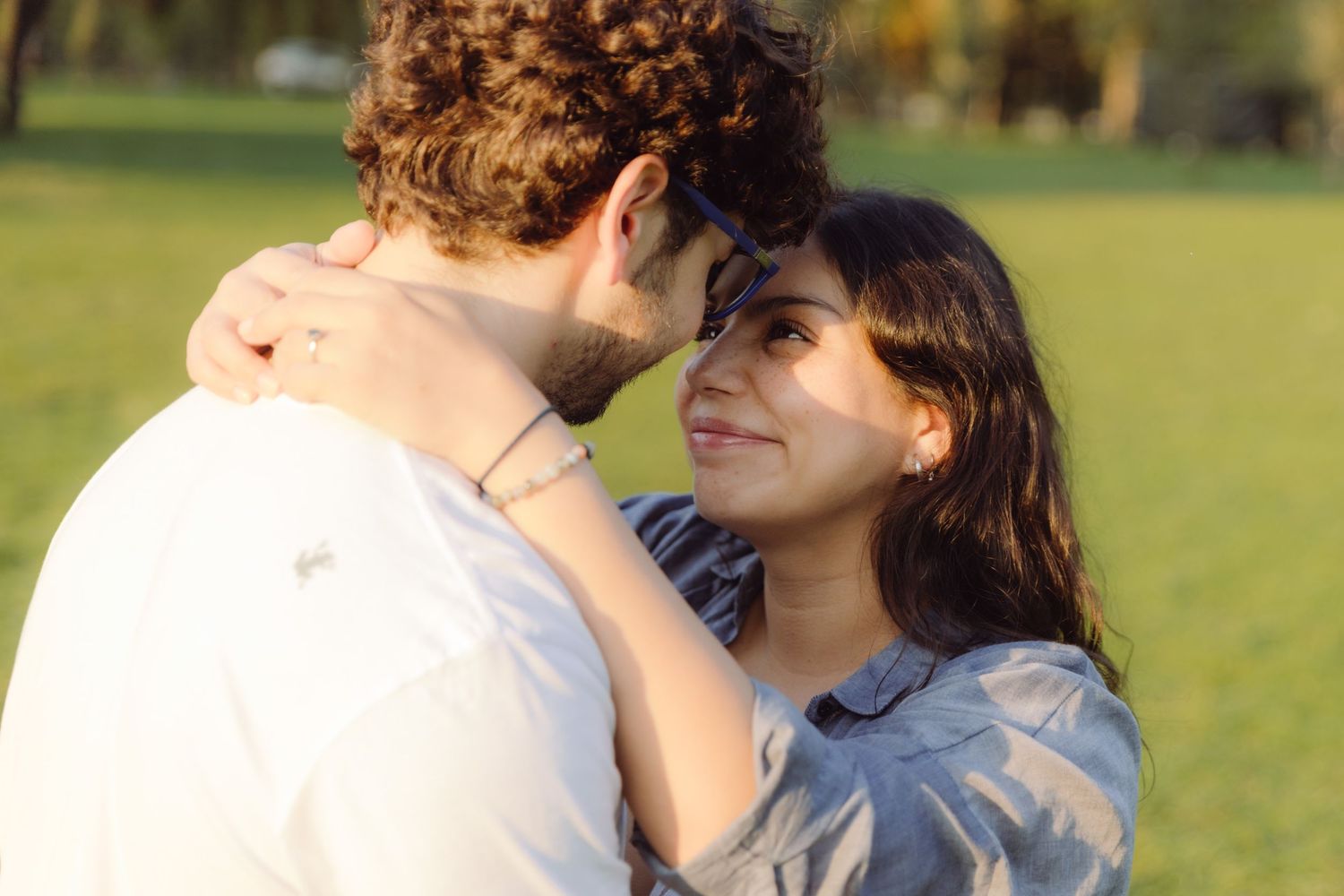 A smiling woman with long dark hair embraces a curly-haired man in a sunny park setting.